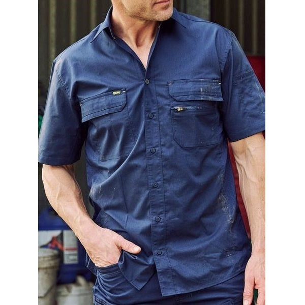 Man wearing a blue work shirt with multiple pockets in an industrial setting.