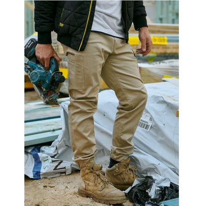 Person wearing beige work pants and brown boots holding a power tool on a construction site.