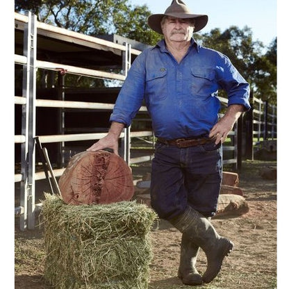 Man in blue work shirt and hat standing next to a hay bale on a farm.