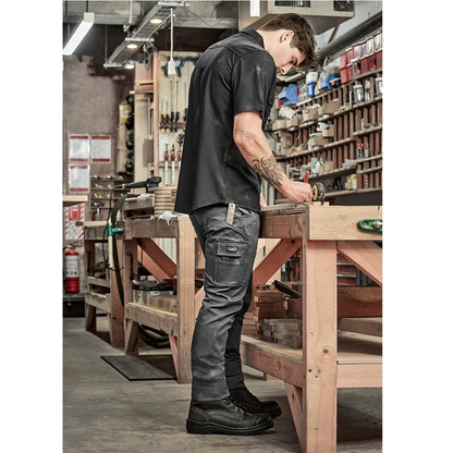 Person working on a wooden bench in a workshop setting with Syzmik ZP230 Men's Essential Basic Stretch Cargo Pants in Charcoal colour