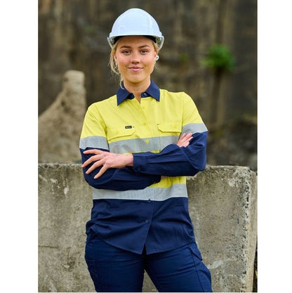 Person wearing a high-visibility work shirt and hard hat standing against a stone wall.